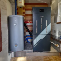 Szary piec Ferroli i zasobnik ciepłej wody Nainei w pomieszczeniu z różnymi rodzajami płytek na ścianach i podłodze, widoczne rury instalacji grzewczej i naczynie przeponowe IBO.