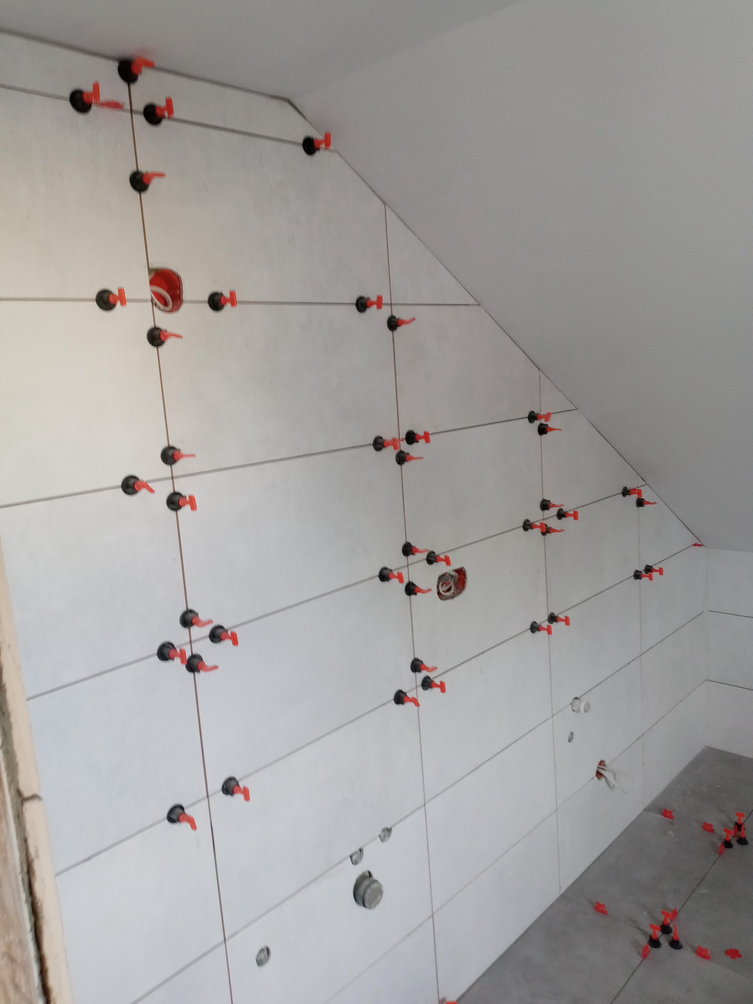 Ściana skośna w trakcie układania jasnych, prostokątnych płytek z użyciem systemu poziomowania z czarnymi podstawkami i czerwonymi klinami, widoczne otwory na instalacje.