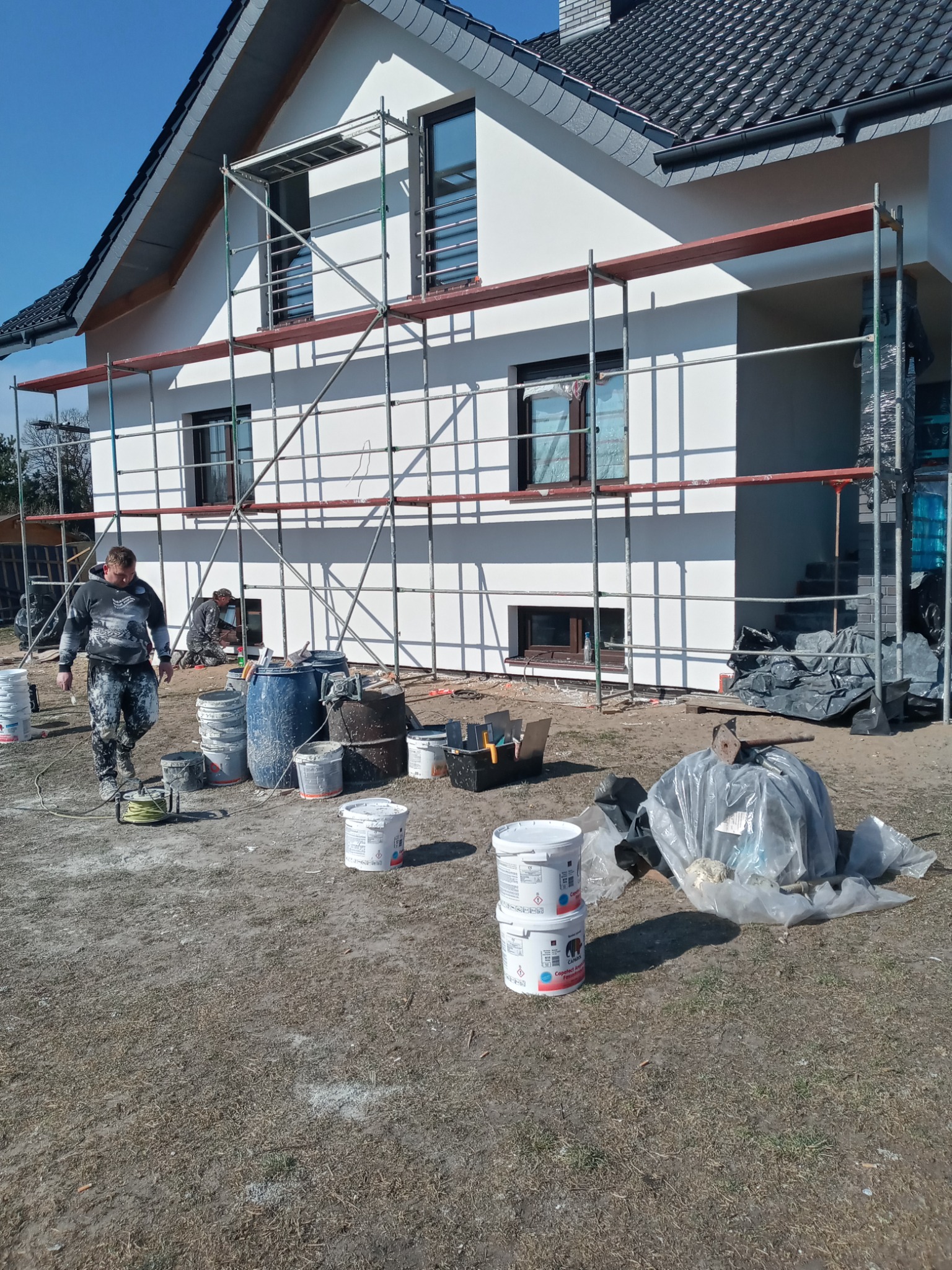 Pracownik w ubrudzonym farbą ubraniu przechodzi obok wiaderek z farbą i rusztowania przy białej elewacji domu jednorodzinnego w trakcie prac wykończeniowych.