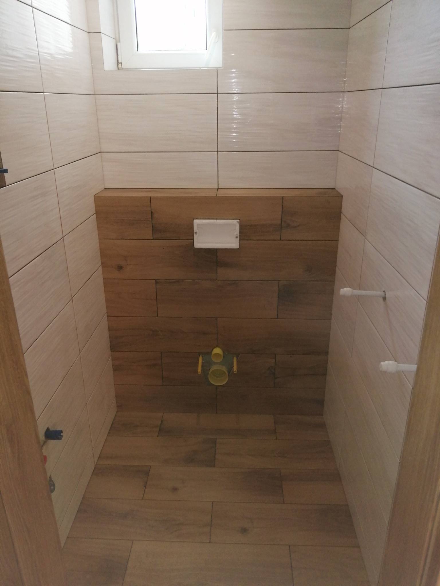 Wnętrze małej łazienki z beżowymi i drewnopodobnymi płytkami, widoczna zabudowa stelaża WC, częściowo ułożona podłoga, przygotowane wyprowadzenia instalacji wodnej.