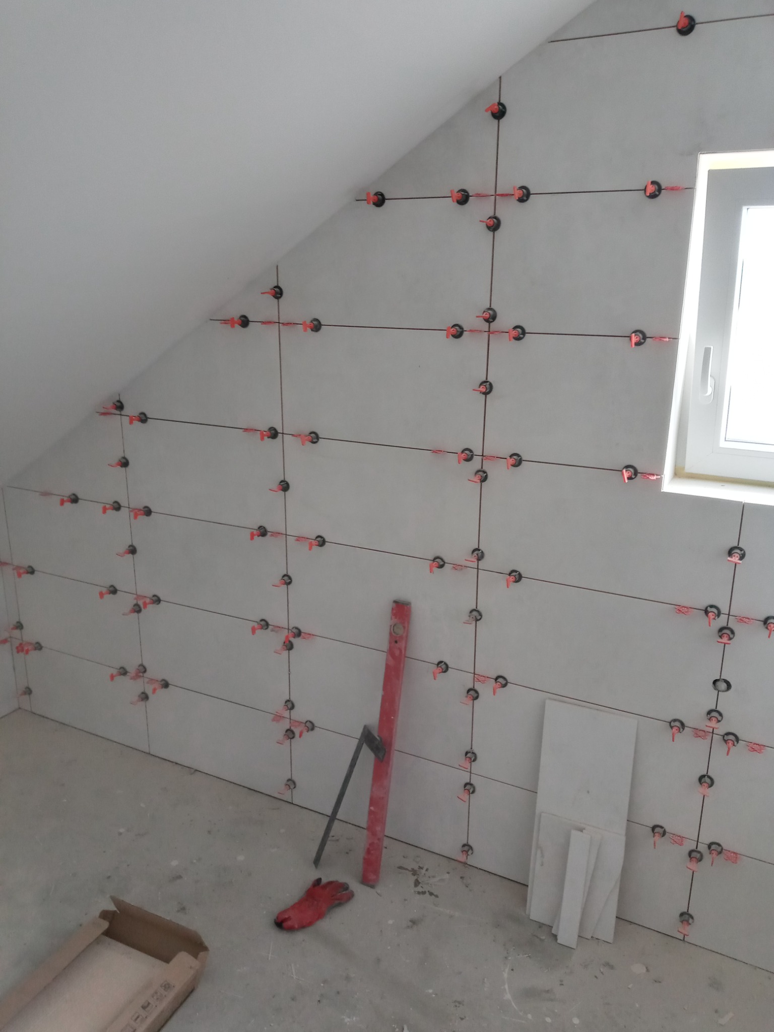 Ściana w skosie pokryta szarymi płytkami z czerwonymi krzyżykami dystansowymi, obok okna, na podłodze karton i rękawica robocza.