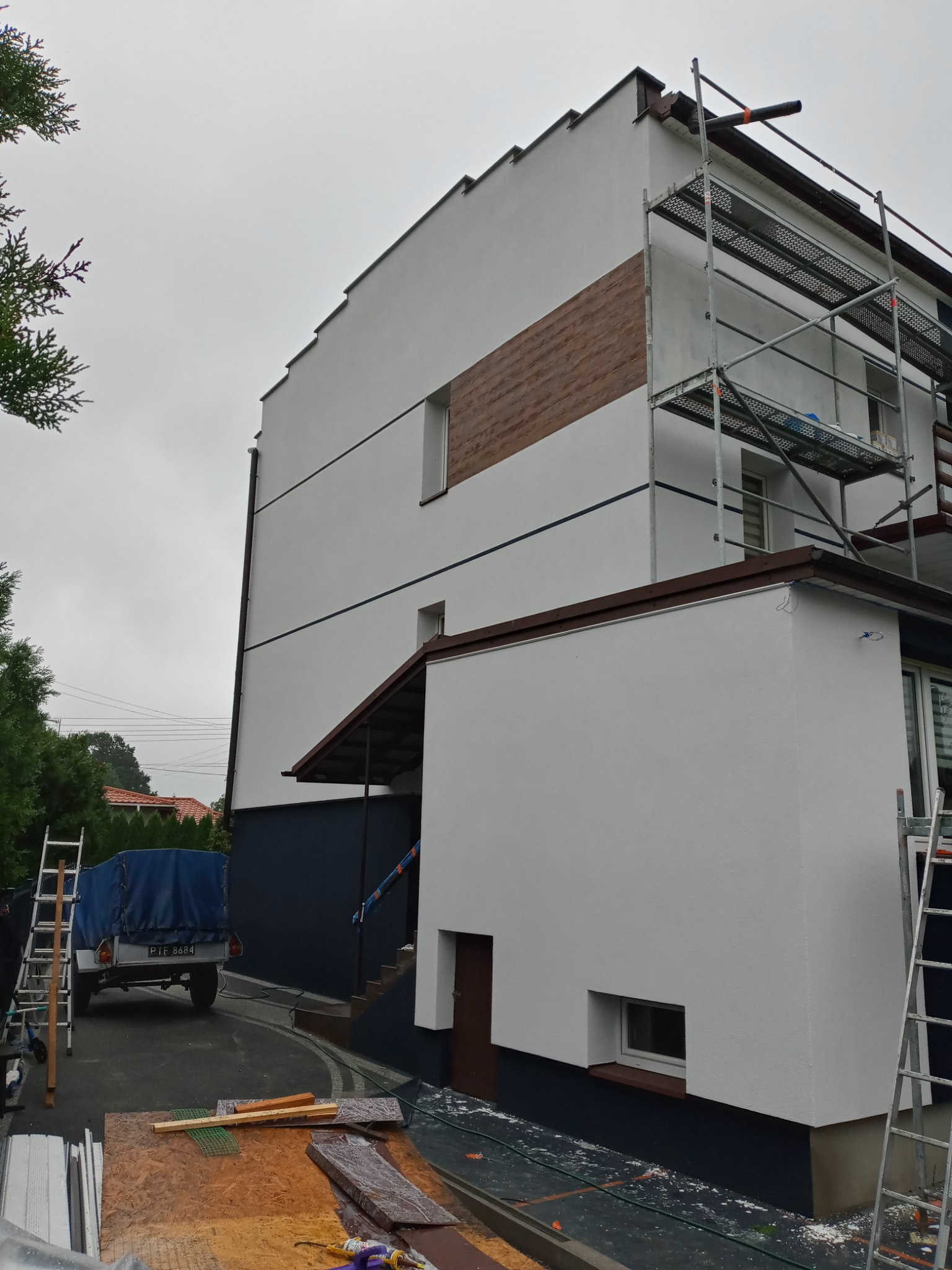 Dwupiętrowy budynek po częściowym ociepleniu elewacji, z widocznym rusztowaniem, przyczepką z plandeką i materiałami budowlanymi na podjeździe w pochmurny dzień.