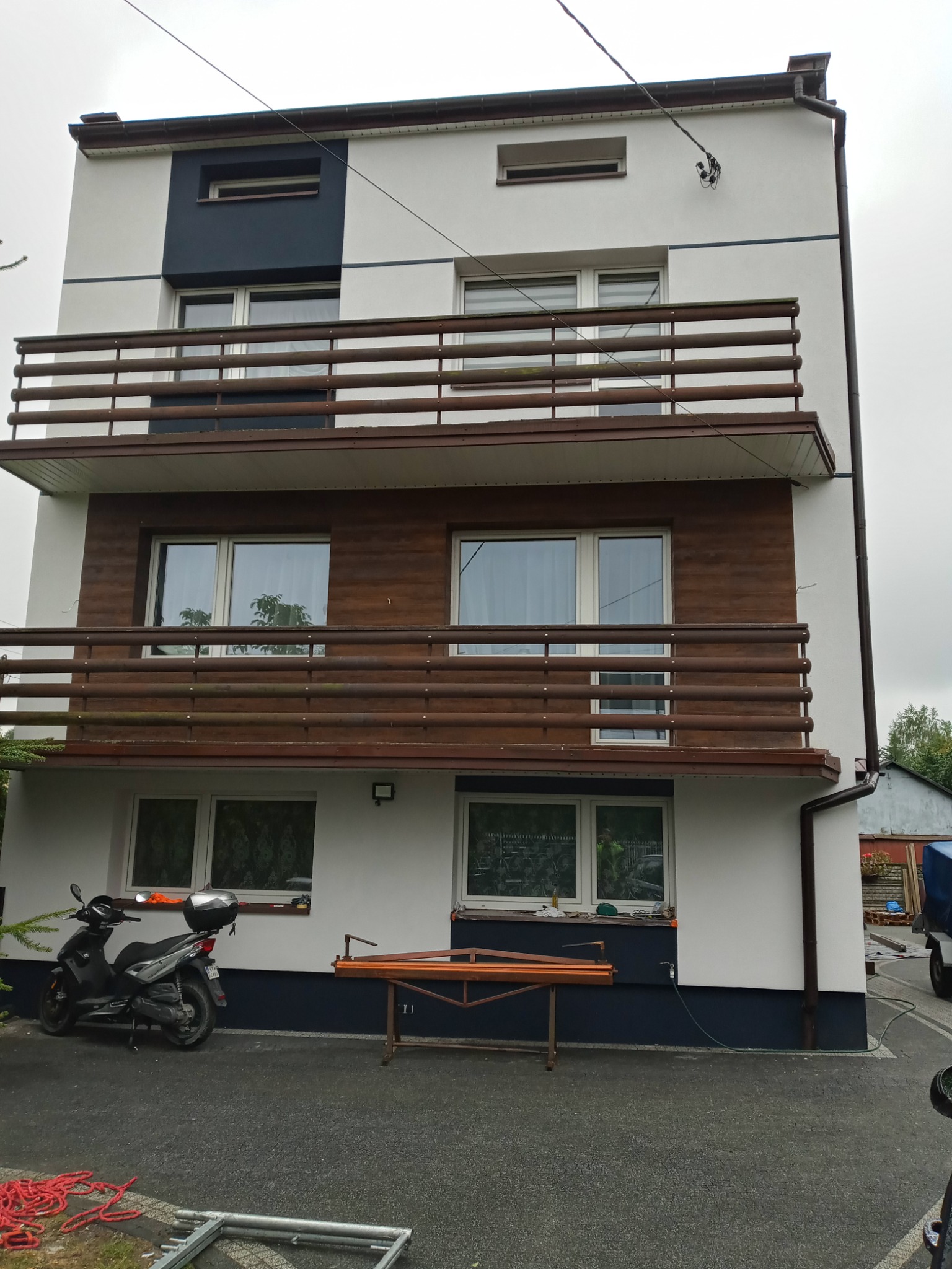 Trzykondygnacyjny dom z balkonami, częściowo wykończony drewnianą okładziną, z motocyklem i narzędziami na podjeździe.