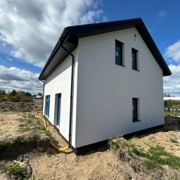 KB Bartłomiej Kowalak - Nowoczesny, biały dom jednorodzinny z czarnym dachem, widoczny z zewnątrz, z żółtą rurą przy fundamencie i niezagospodarowanym terenem wokół. Błękitne niebo z chmurami w tle.