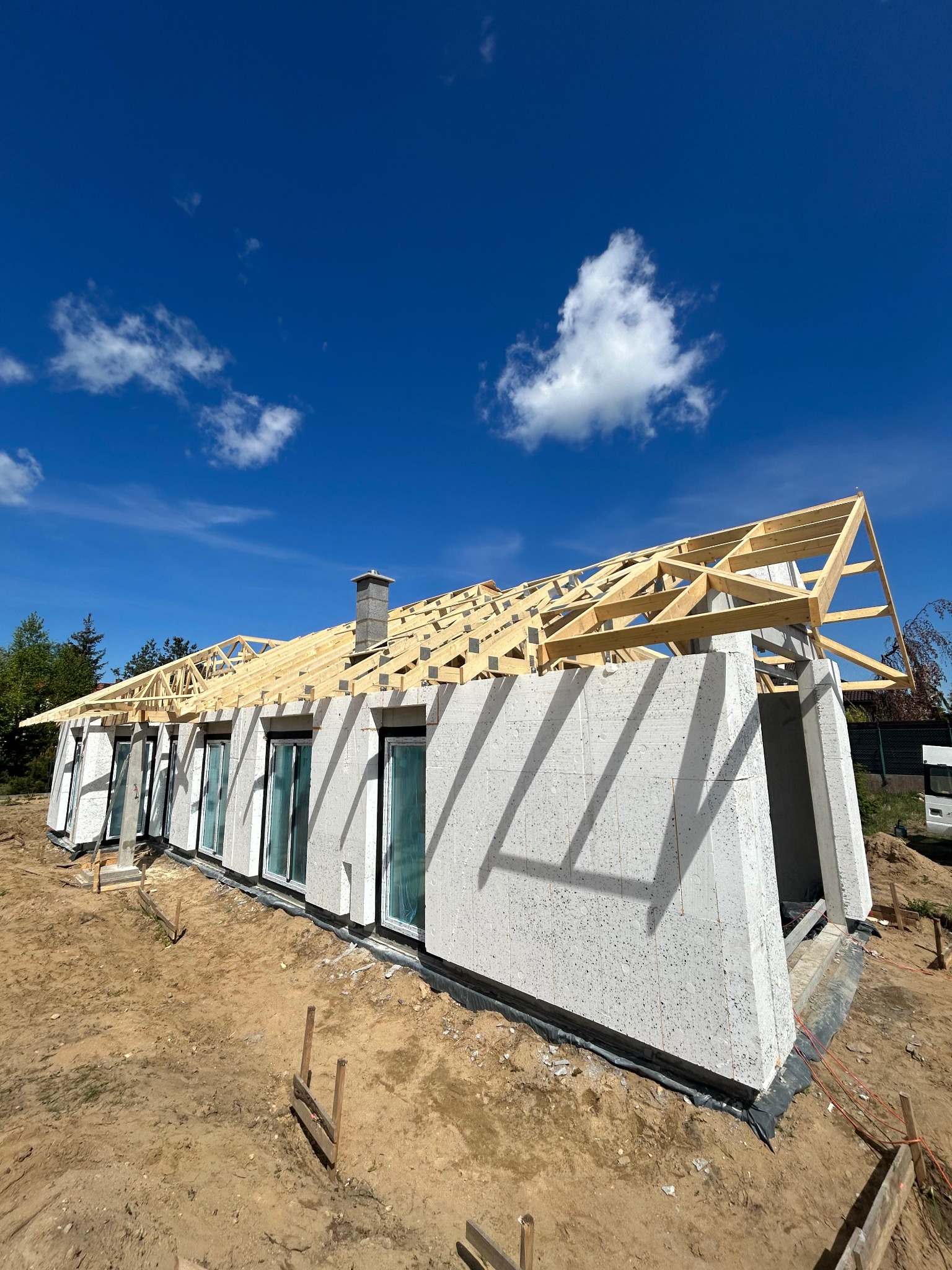 Budowa domu jednorodzinnego z betonu komórkowego: ściany, okna, więźba dachowa w trakcie montażu, komin. Widok z zewnątrz, słoneczny dzień.