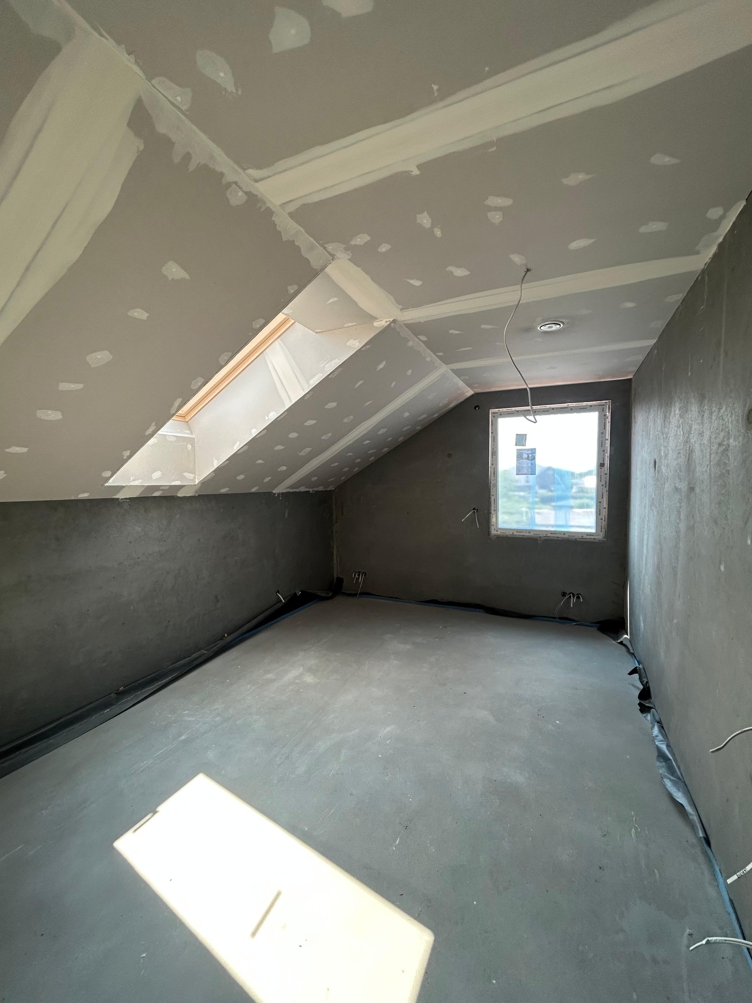 Poddasze w trakcie wykańczania: surowe ściany, okno dachowe i wylewka. Widoczne przewody elektryczne i przygotowanie do dalszych prac wykończeniowych.