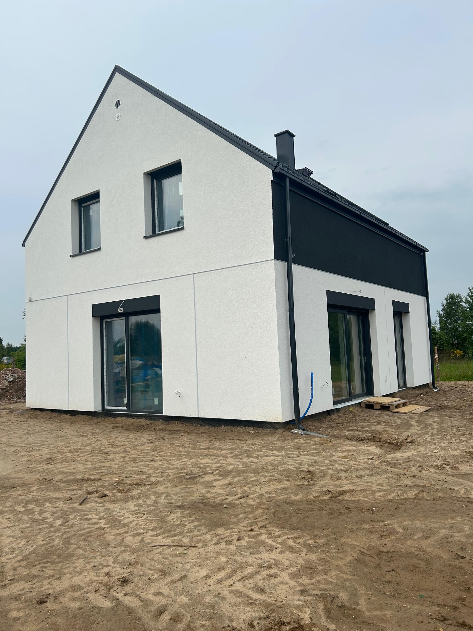 Nowoczesny, dwukondygnacyjny dom z białymi ścianami i czarnymi akcentami, w trakcie wykończenia. Widoczne okna, drzwi przesuwne i teren wokół budynku.
