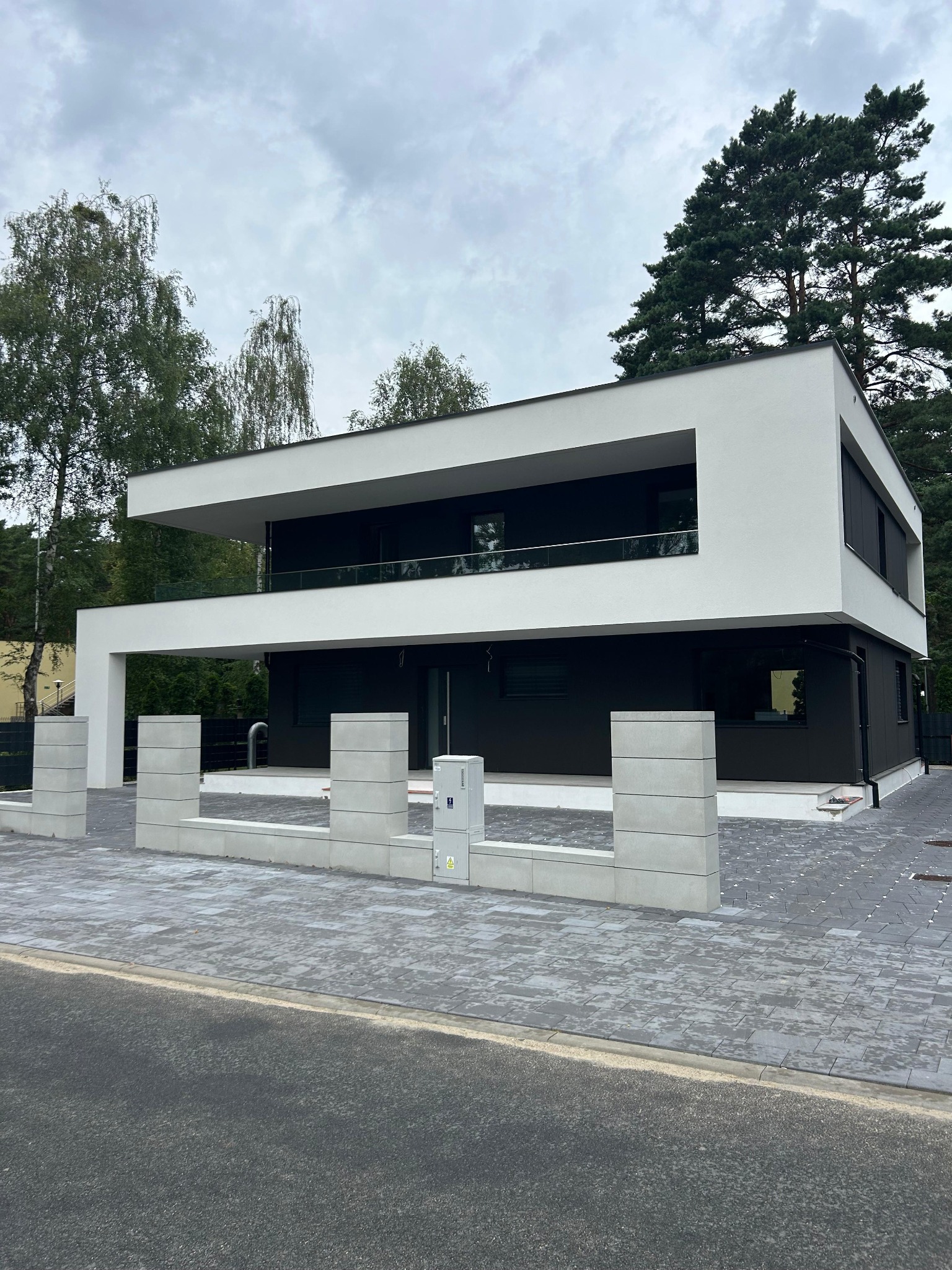 Dwukondygnacyjny dom w stylu nowoczesnym z płaskim dachem, białą elewacją i czarnymi panelami, otoczony zielenią. Widoczny fragment chodnika z kostki brukowej.