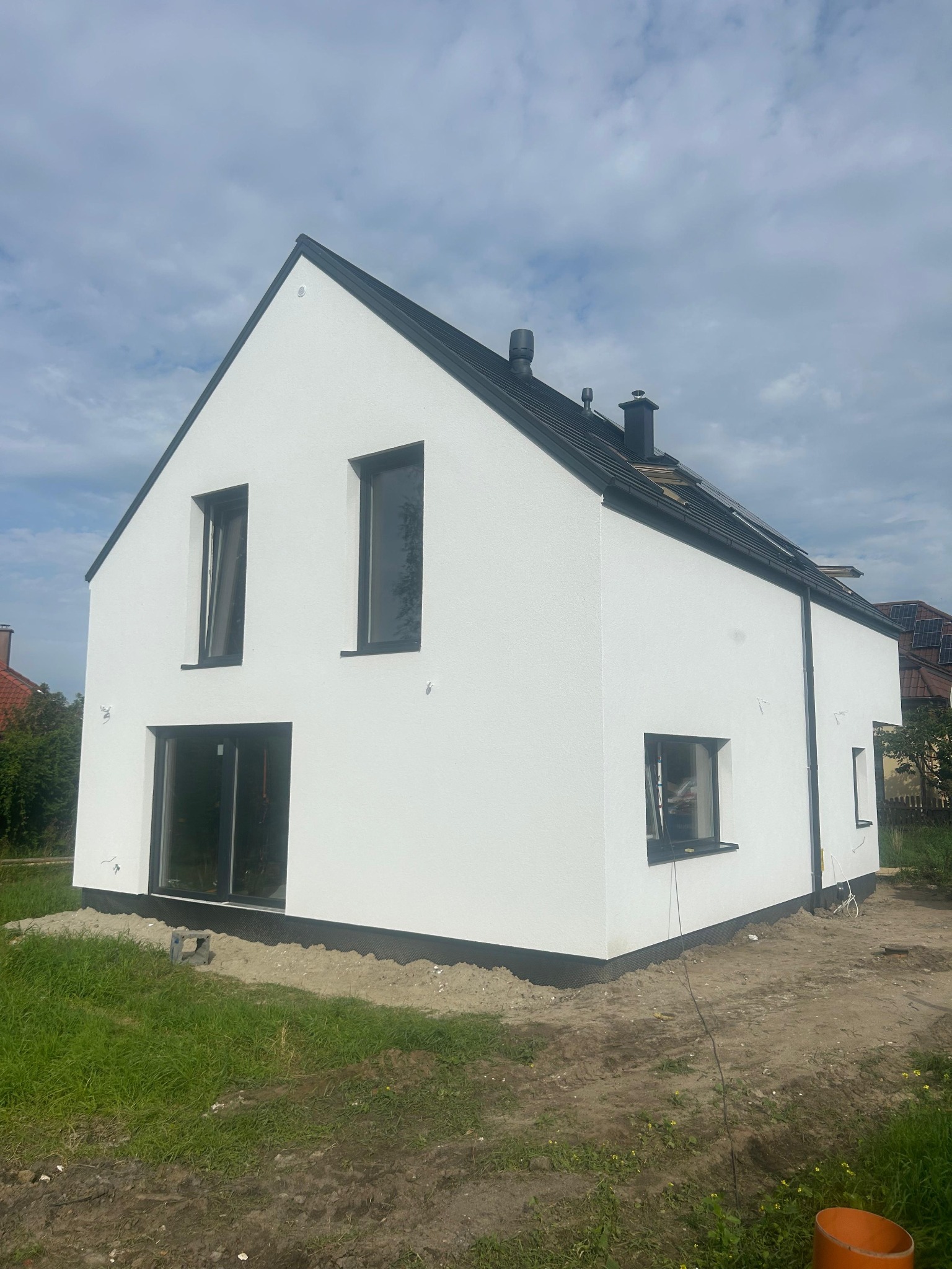 Nowoczesny, biały dom jednorodzinny z czarną stolarką okienną i dachem, w trakcie wykończenia elewacji. Widoczne fragmenty instalacji na dachu i otoczenie w stanie surowym.