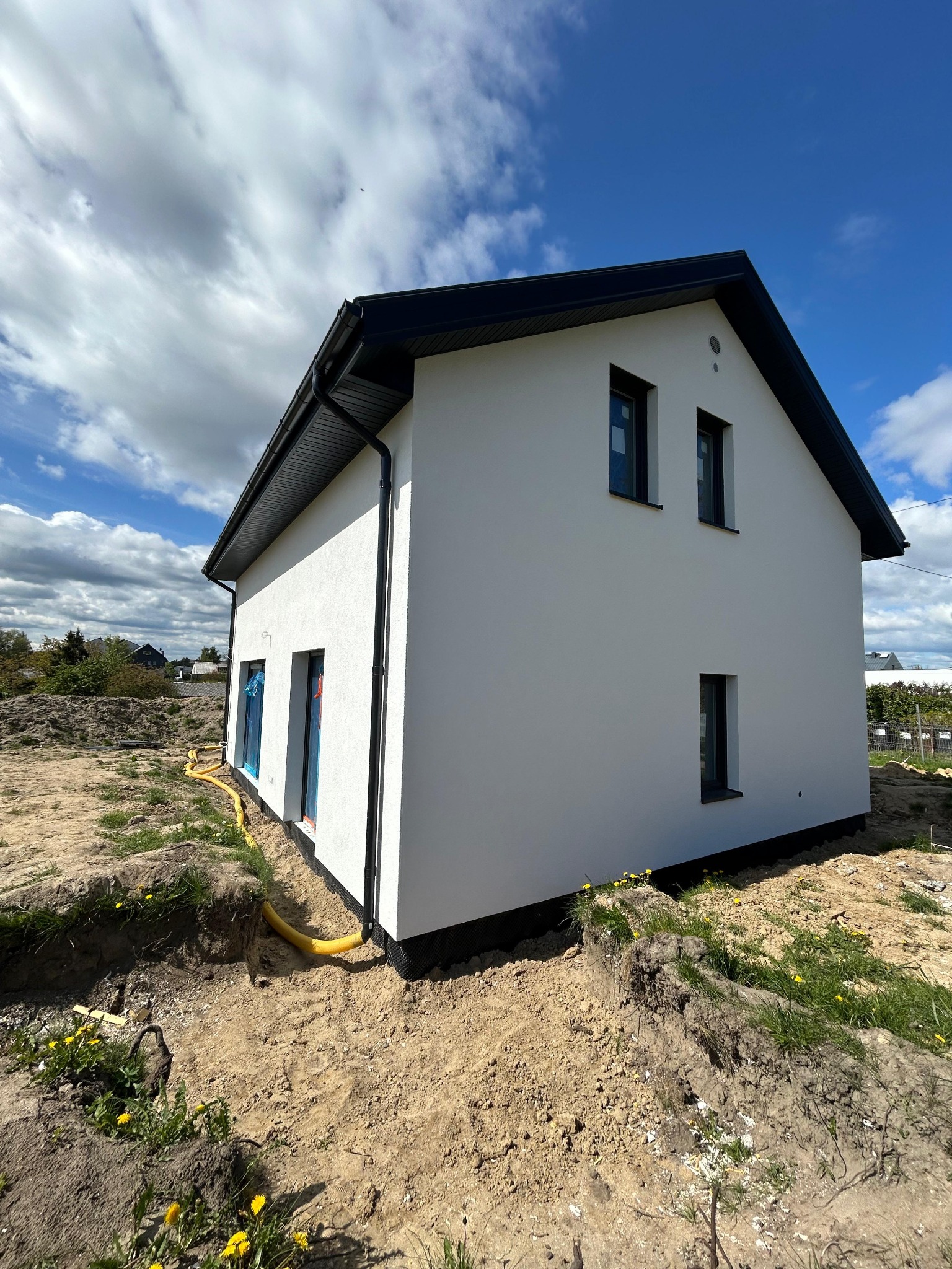 Nowoczesny, biały dom jednorodzinny z czarnym dachem, widoczny z zewnątrz, z żółtą rurą przy fundamencie i niezagospodarowanym terenem wokół. Błękitne niebo z chmurami w tle.