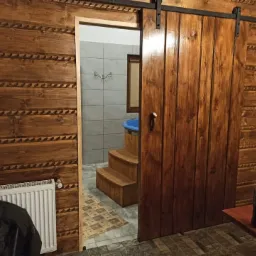 Wnętrze rustykalnego pokoju z drewnianymi ścianami i przesuwnymi drzwiami do łazienki z drewnianą wanną i schodkami.