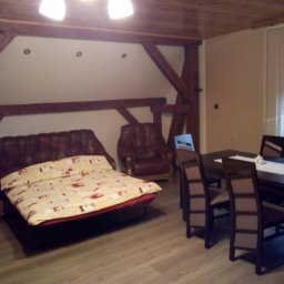 PPHU ROLMIX - Wnętrze pokoju z drewnianą konstrukcją stropu, rozkładaną sofą z narzutą w geometryczne wzory, skórzanym fotelem, stołem z krzesłami i butelkami wody mineralnej.