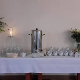 Stół z białym obrusem przygotowany do serwowania herbaty, z dekoracjami kwiatowymi, świecznikami i srebrnym zaparzaczem do wody.