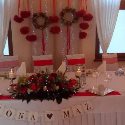 Udekorowany st&oacute;ł weselny z napisem 'Żona Mąż' na tle białych zasłon i czerwonych ozd&oacute;b, świece w szklanych świecznikach, czerwono-białe dekoracje krzeseł.