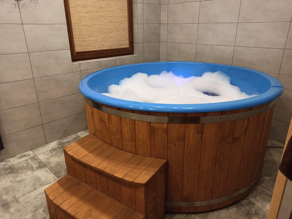 Drewniana beczka kąpielowa z niebieskim wnętrzem wypełniona pianą, z drewnianymi schodkami, ustawiona w pomieszczeniu z szarymi płytkami.