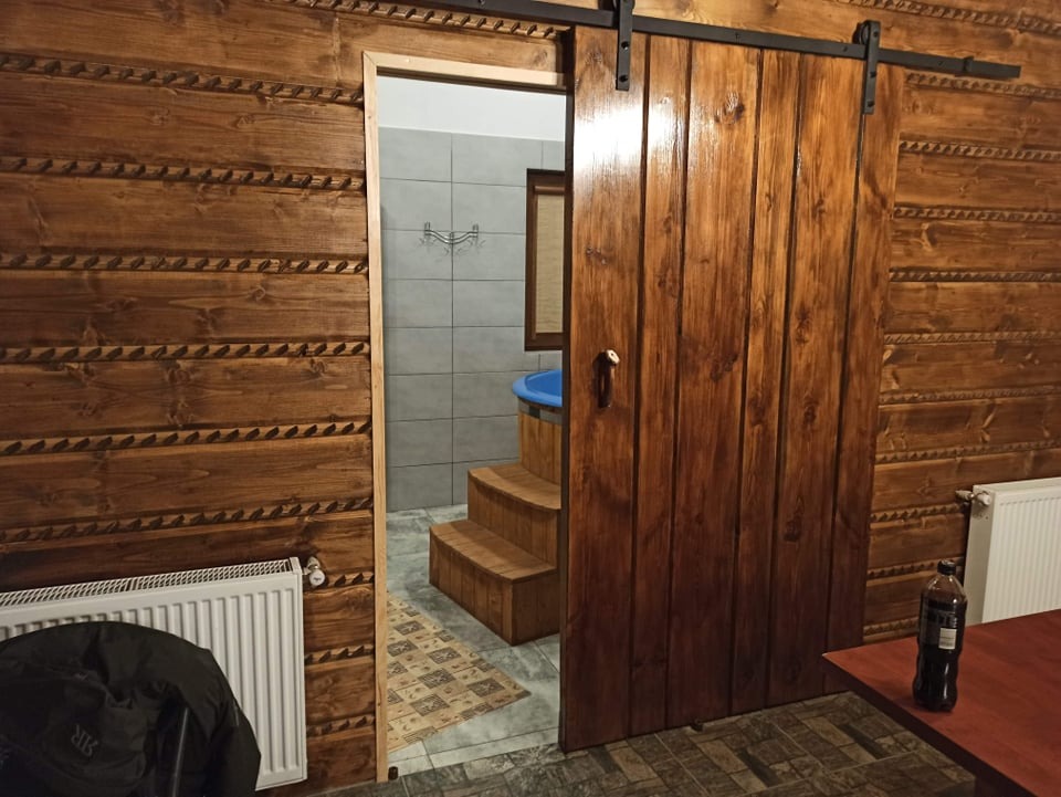 Wnętrze rustykalnego pokoju z drewnianymi ścianami i przesuwnymi drzwiami do łazienki z drewnianą wanną i schodkami.