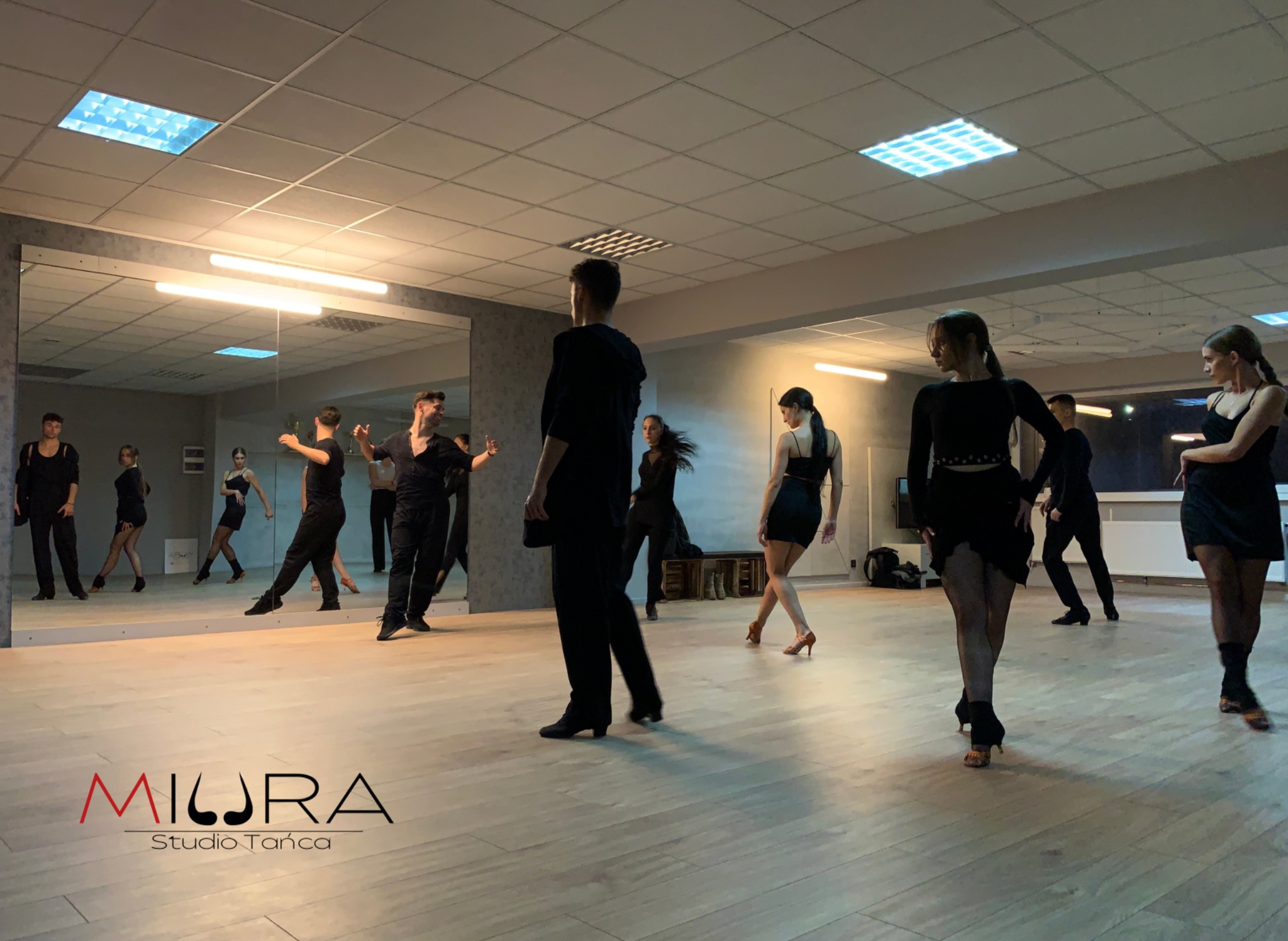 Grupa osób w czarnych strojach ćwiczy taniec w sali z lustrami, widoczne logo studia tańca w lewym dolnym rogu.