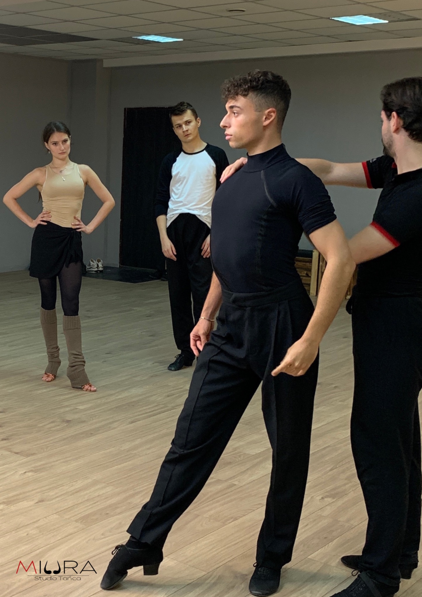 Instruktor koryguje postawę tancerza podczas lekcji, w tle obserwują dwie osoby.