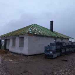 Budowa domu jednorodzinnego w stanie surowym otwartym, widoczna drewniana konstrukcja dachu bez pokrycia, palety z dachówką Creaton obok budynku