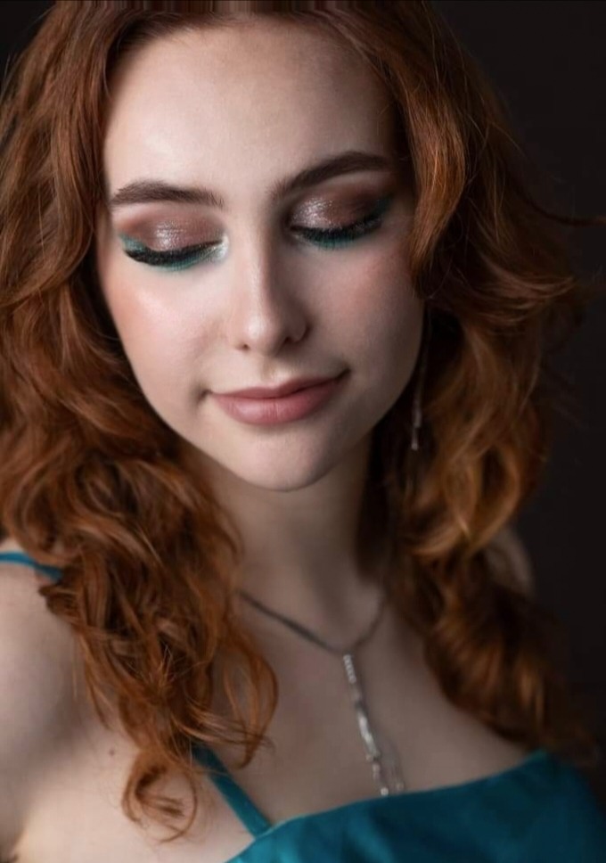 Portret młodej kobiety z rudymi, kręconymi włosami i delikatnym, błyszczącym eye-art w odcieniach brązu i turkusu, z subtelnym uśmiechem.