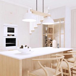 Decoria Studio Patrycja Tachmańska - Jasna kuchnia z wyspą, drewnianymi hokerami i zabudową meblową z piekarnikiem; nad wyspą wiszą dwie białe lampy z drewnianym elementem mocującym, w tle widoczna ażurowa półka i schody.