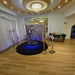 Okrągła platforma z niebieskim podświetleniem, automat do robienia zdjęć i błyszczące tło w sali z dekoracyjnym sufitem i rekwizytami do zdjęć.