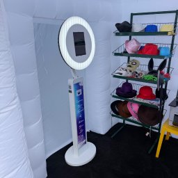 PSOCIMY ANETA DOBRZYŃSKA - Nowoczesne stanowisko do robienia zdjęć z ring lightem w białej, dmuchanej kabinie, obok zielony regał z różnorodnymi rekwizytami: kapeluszami, maskami i zabawnymi akcesoriami.