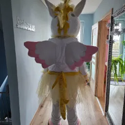 Osoba w kostiumie białego jednorożca z różowymi skrzydłami i złotą grzywą, widziana od tyłu w korytarzu mieszkania.