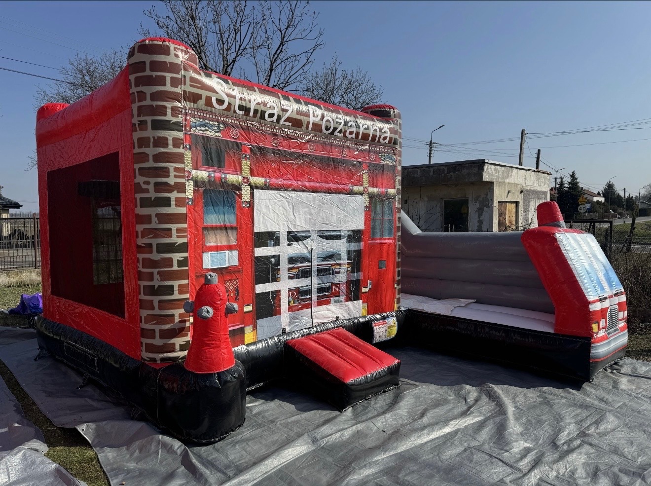 Duży, czerwony dmuchany zamek w kształcie remizy strażackiej z napisem 'Straż Pożarna', hydrantem i elementami imitującymi cegły, ustawiony na szarej plandece.