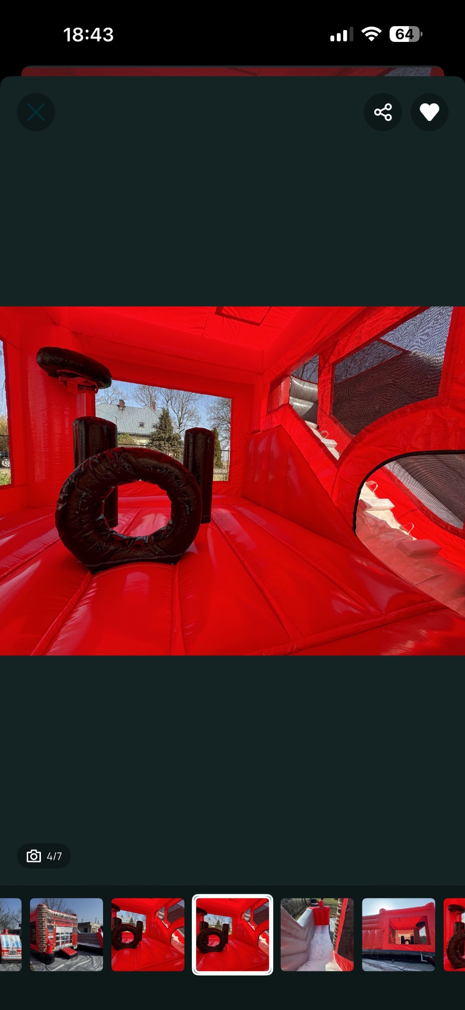 Czerwony dmuchany zamek z przeszkodami wewnątrz, widok na zewnątrz przez okno, czarne, okrągłe przeszkody na podłodze. Jasne oświetlenie.