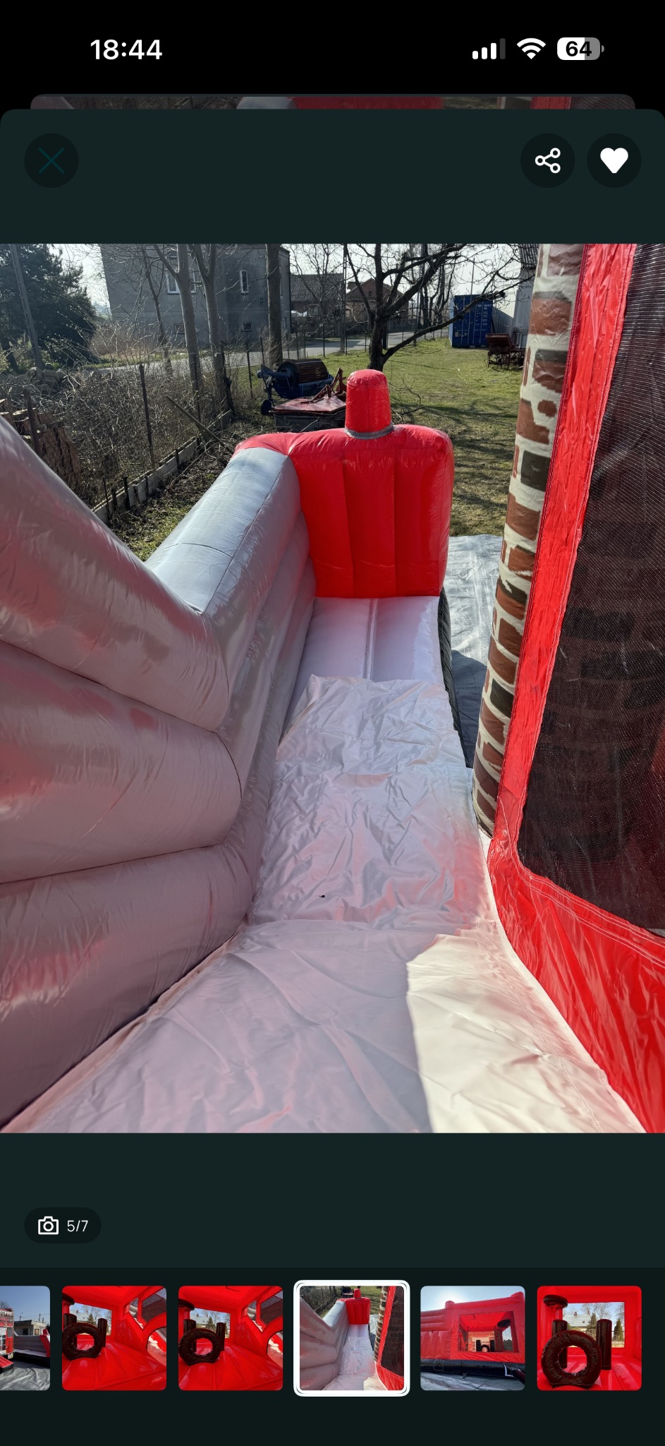 Wnętrze dmuchanej zjeżdżalni w kolorach czerwieni i bieli, widok z góry na ślizg. W tle budynek i drzewa. Dmuchaniec ustawiony na trawie.