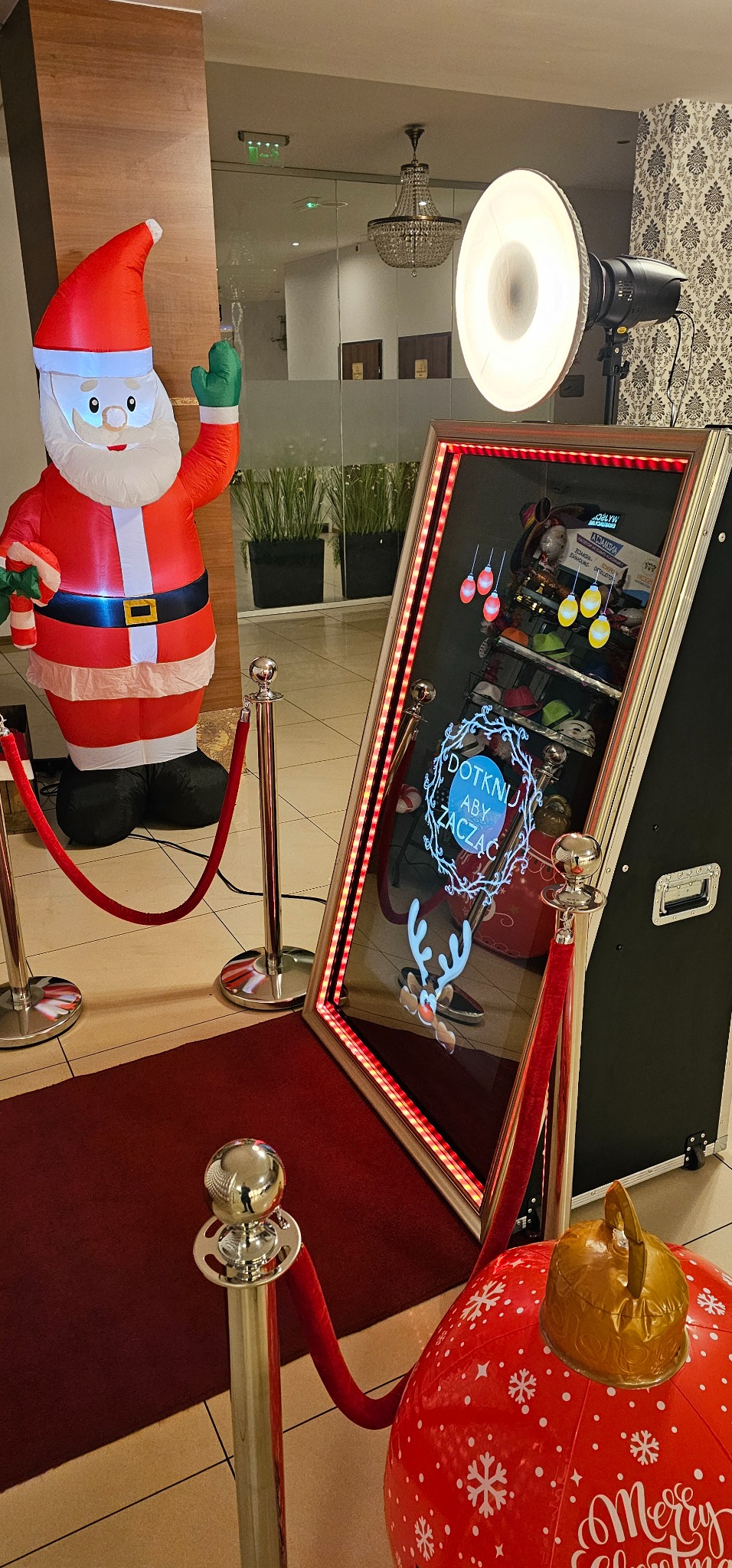 Świąteczna konfiguracja z dmuchanym Świętym Mikołajem, dekoracyjną bombką i lustrzaną kabiną z napisem 'Dotknij aby zacząć', oświetloną lampą błyskową i odgrodzoną czerwonymi sznurami.