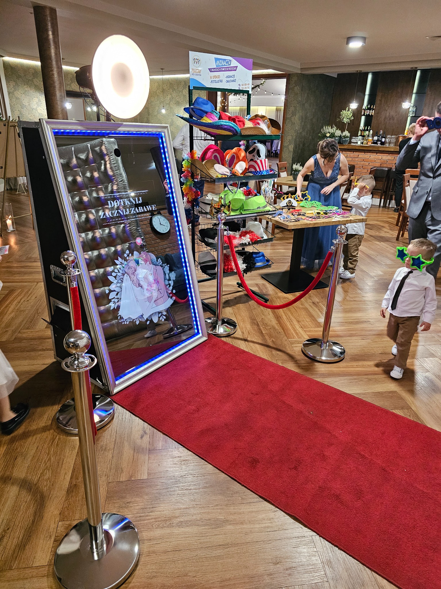 Interaktywne lustro z niebieskim podświetleniem na czerwonym dywanie, obok stolik z rekwizytami do zdjęć i bawiące się dzieci z dorosłą osobą w tle.