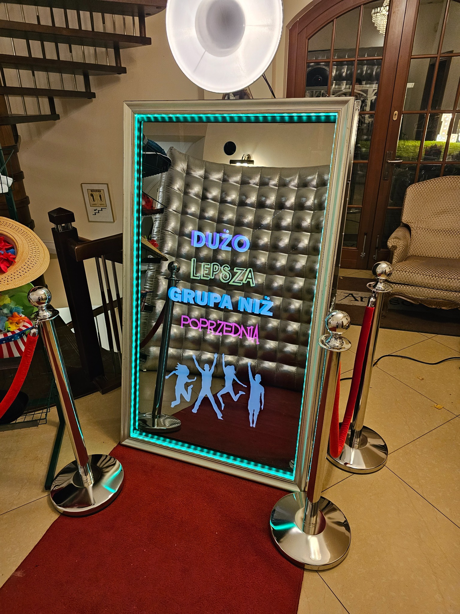 Lustrzana kabina fotograficzna z podświetleniem LED i napisem 'Dużo lepsza grupa niż poprzednia', otoczona słupkami z czerwonymi sznurami na czerwonym dywanie.