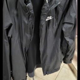 Tekstylowe love - Czarna, rozpięta kurtka sportowa marki Nike z kapturem, wisząca na niebieskim wieszaku, widoczne logo Nike na piersi, w tle inne ubrania na wieszakach.