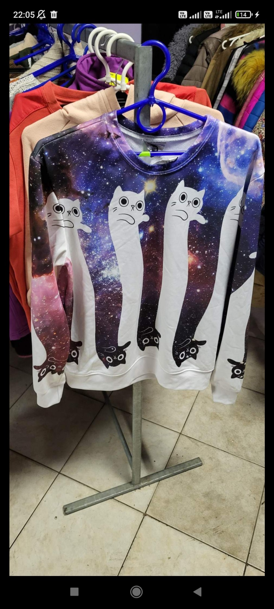 Bluza z nadrukiem kosmicznego tła i stylizowanych, białych kotów z długimi szyjami, wisząca na wieszaku.