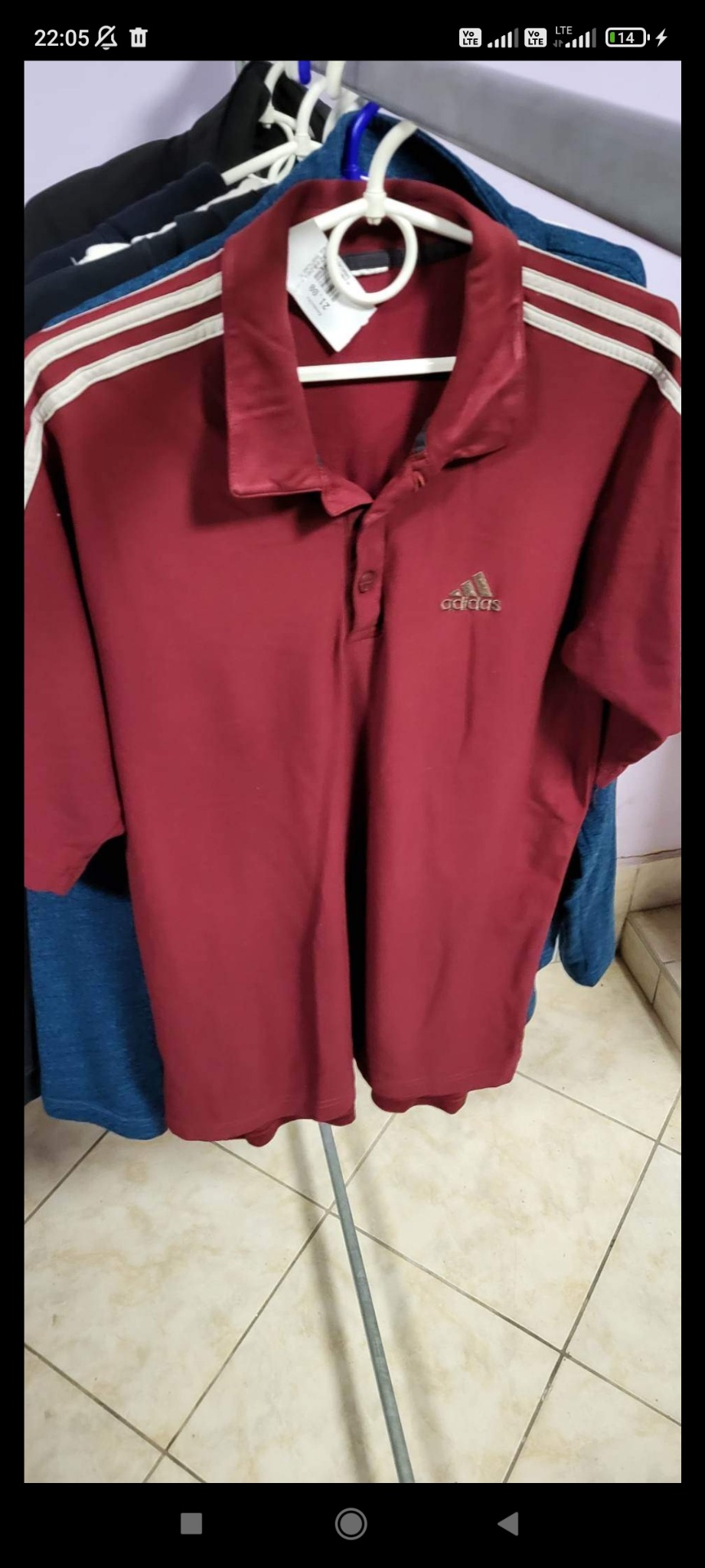 Bordowa koszulka polo marki Adidas z białymi paskami na ramionach, wisząca na wieszaku, z widoczną metką i logo firmy na tle innych ubrań.