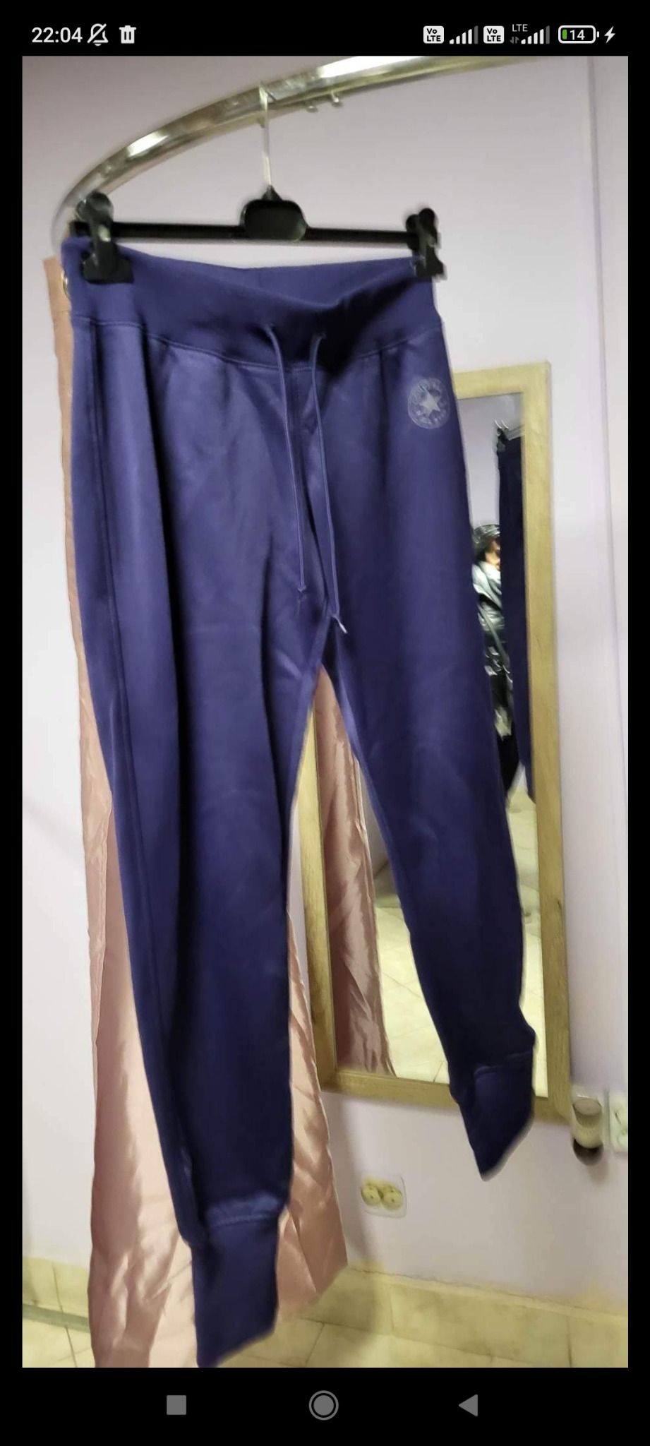 Granatowe spodnie dresowe marki Converse z widocznym logo na nogawce, wiszące na czarnym wieszaku na tle lustra i beżowej zasłony.