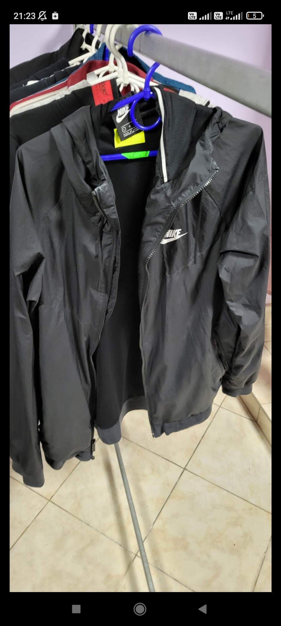 Czarna, rozpięta kurtka sportowa marki Nike z kapturem, wisząca na niebieskim wieszaku, widoczne logo Nike na piersi, w tle inne ubrania na wieszakach.