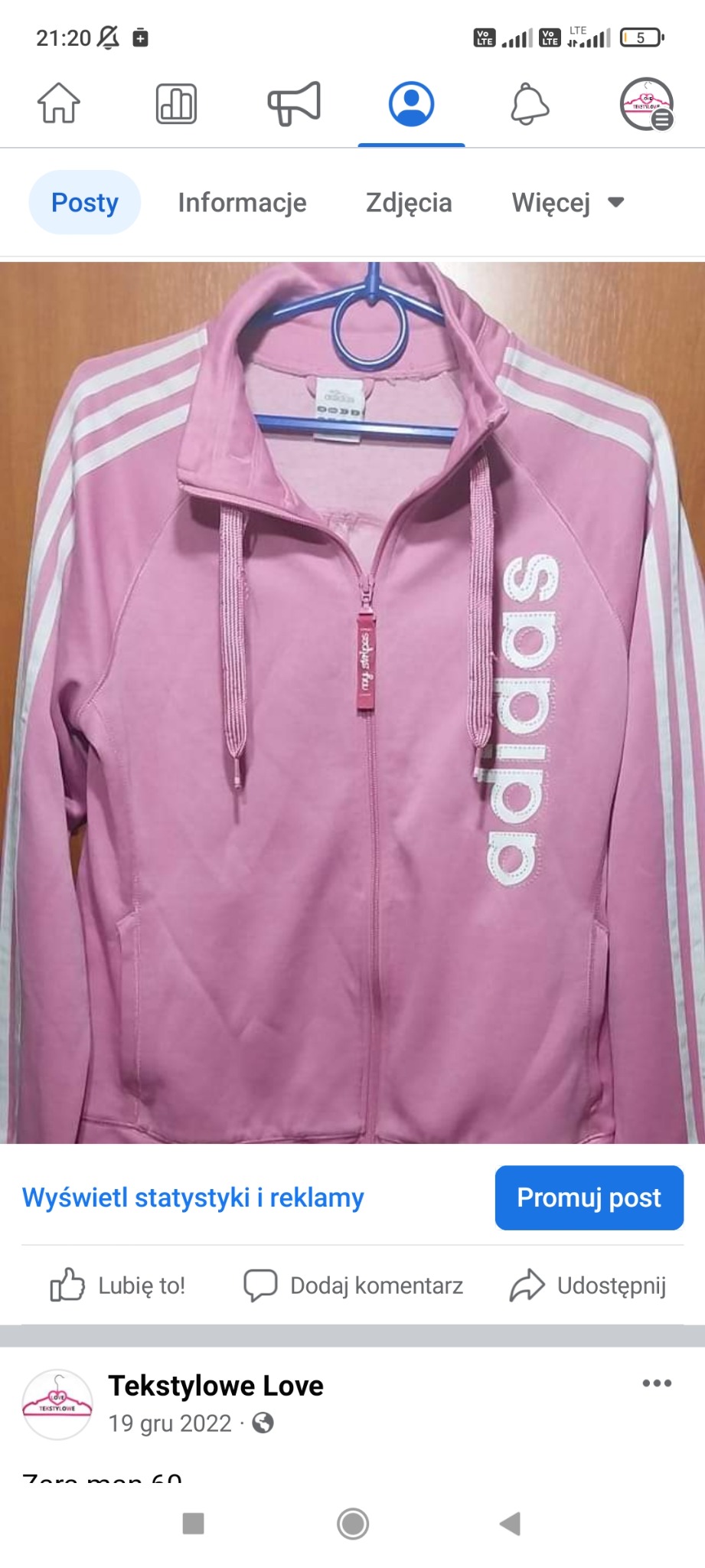 Różowa bluza sportowa Adidas z białymi paskami na ramionach, wisząca na niebieskim wieszaku, z widocznym suwakiem i napisem 'adidas' na boku.