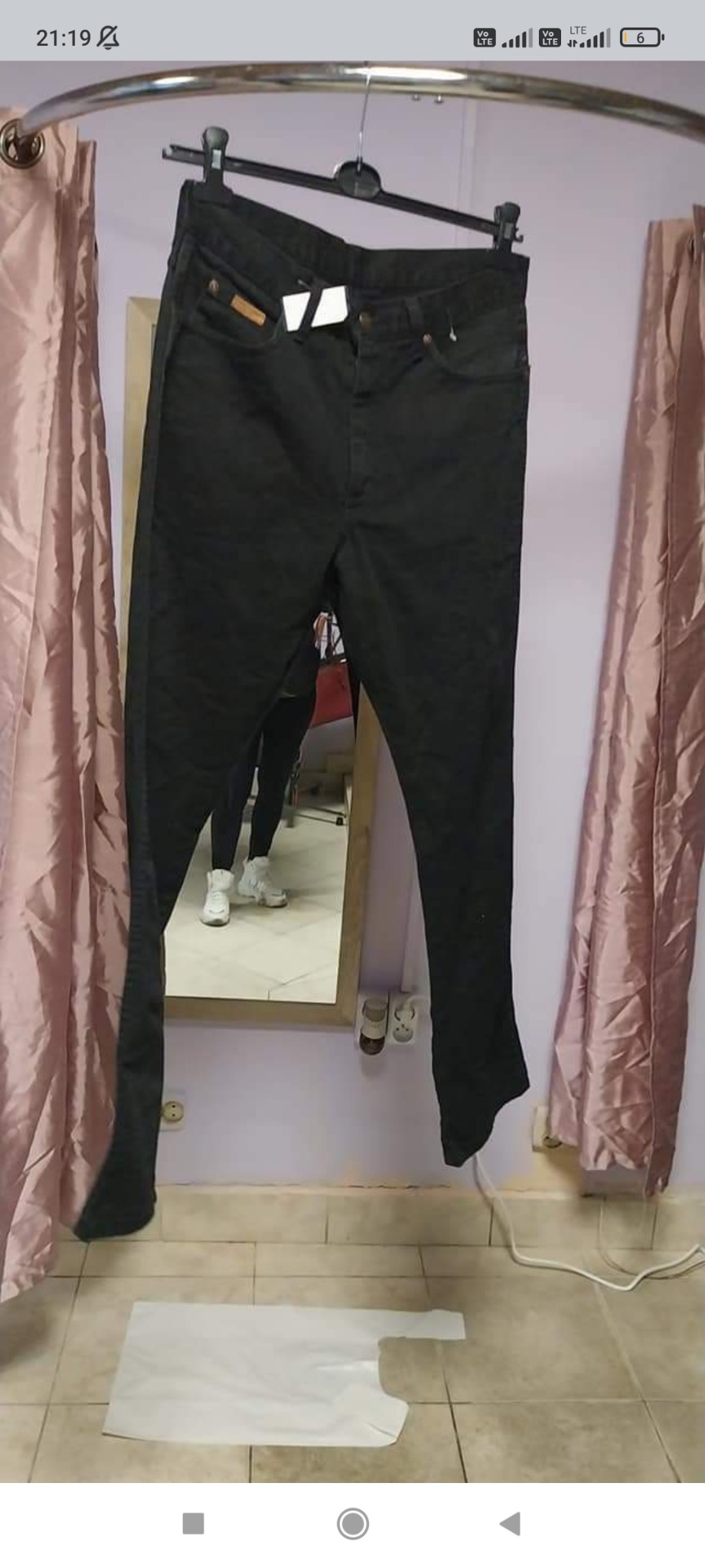 Czarne spodnie jeansowe z metką, wiszące na wieszaku w przymierzalni z lustrem, w którym odbija się osoba w białych butach. Na podłodze leży biała torba plastikowa.