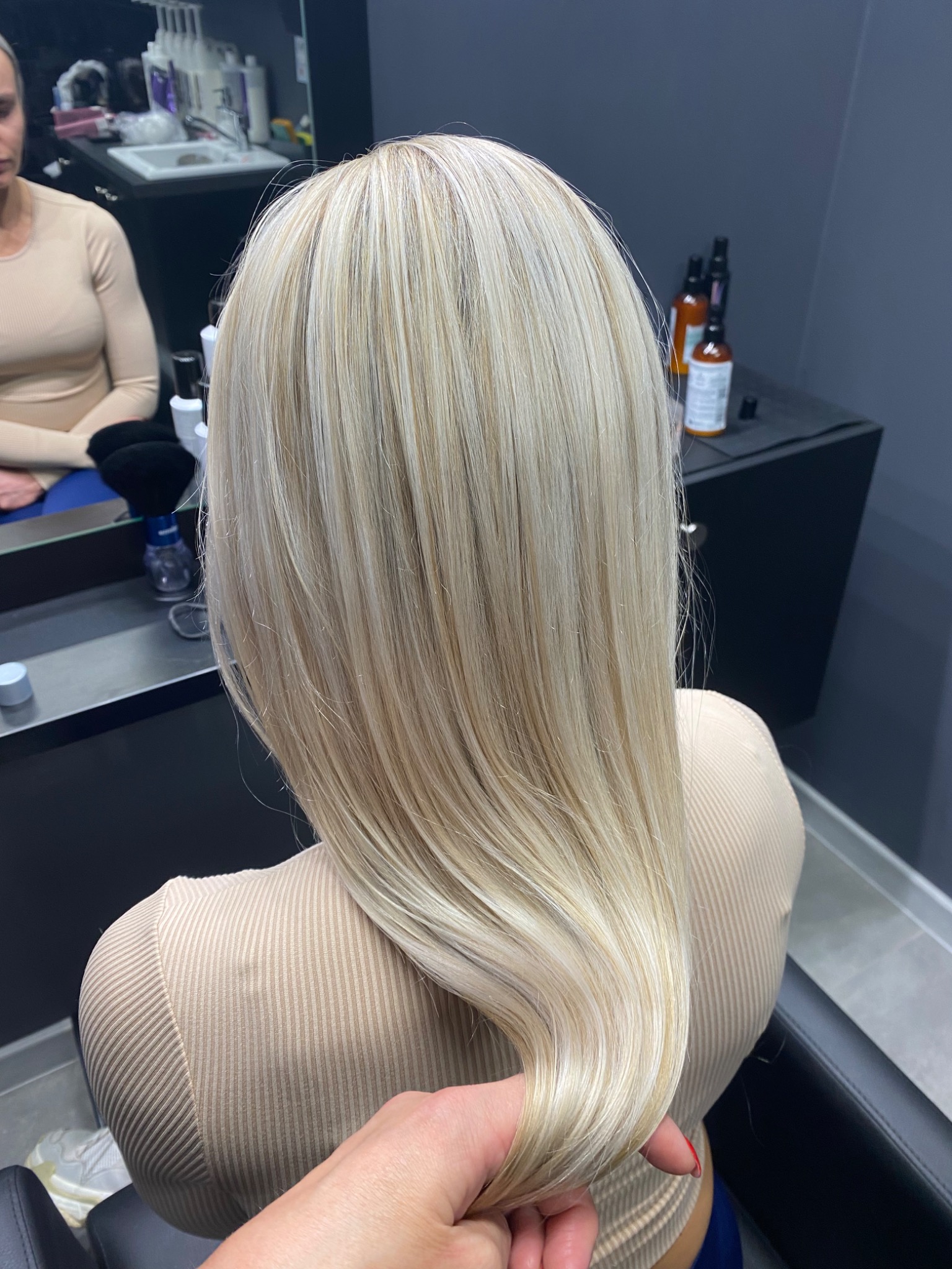 Tył głowy kobiety z długimi, prostymi, blond włosami po koloryzacji z widoczną dłonią fryzjera sprawdzającą teksturę włosów, w tle lustro i kosmetyki.