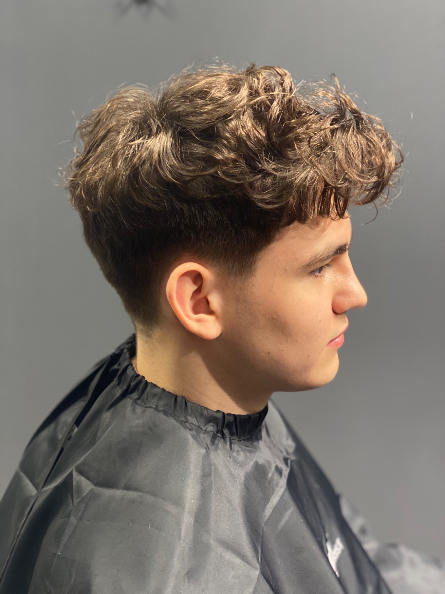 Profil młodego mężczyzny z kręconymi, brązowymi włosami po strzyżeniu, widoczny w czarnej pelerynie ochronnej na tle szarej ściany.