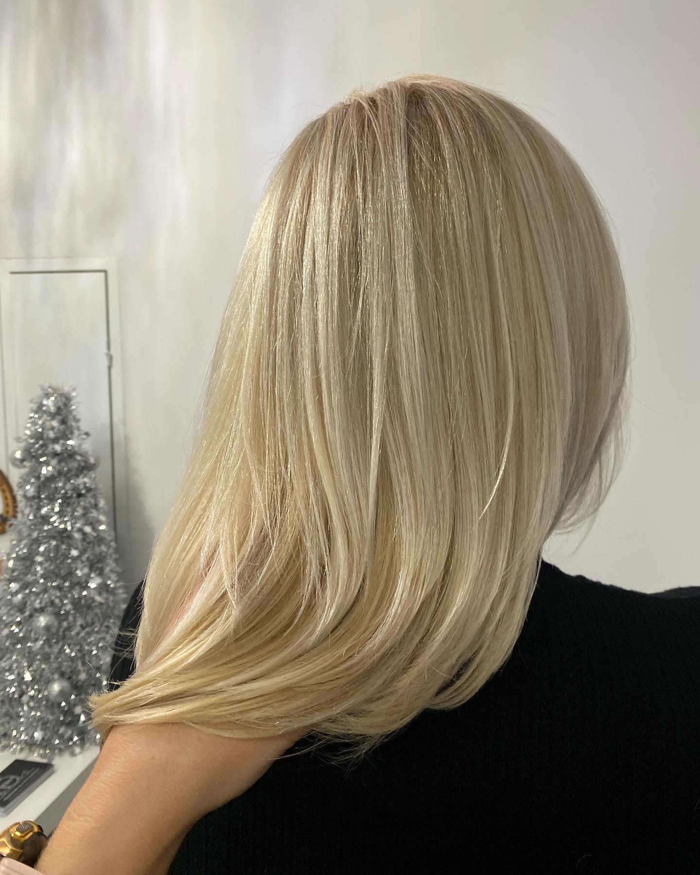 Świeżo ostrzyżone, platynowe blond włosy do ramion, widok z tyłu, delikatny skręt końcówek, w tle srebrna choinka i drzwi.