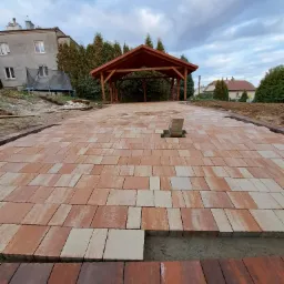 Nowo ułożona, wzorzysta nawierzchnia z kostki w odcieniach czerwieni i beżu, z widocznym fragmentem niedokończonego obrzeża i stertą kostek na środku, drewniana altana w tle.