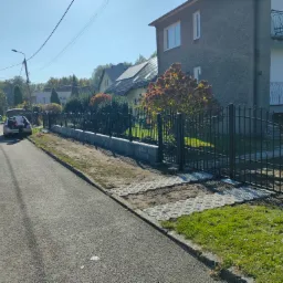 Nowoczesne, czarne ogrodzenie panelowe z bramą przesuwną, zamontowane na betonowej podmur&oacute;wce, widoczne od strony ulicy, z samochodem i osobą przy nim. Na posesji widoczny dom z balkonem i krzewy.