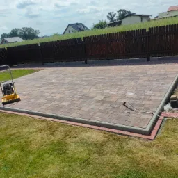 Ułożona kostka brukowa w odcieniach brązu i szarości, obrzeża z czerwonej kostki, ubijarka wibracyjna na powierzchni, widok z góry na trawnik i ogrodzenie.