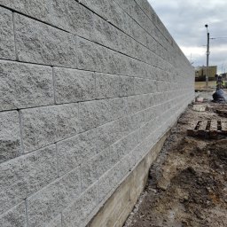 OGROBUD - Szara ściana oporowa z betonowych bloczków, widoczna z boku, z fundamentem i ziemią u podstawy. W tle niebo i budynki.