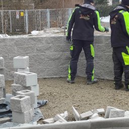 OGROBUD - Budowa muru z szarej kostki betonowej. Dwóch pracowników w odzieży roboczej z neonowymi wstawkami. Częściowo ukończony mur i przygotowane elementy do dalszej budowy.