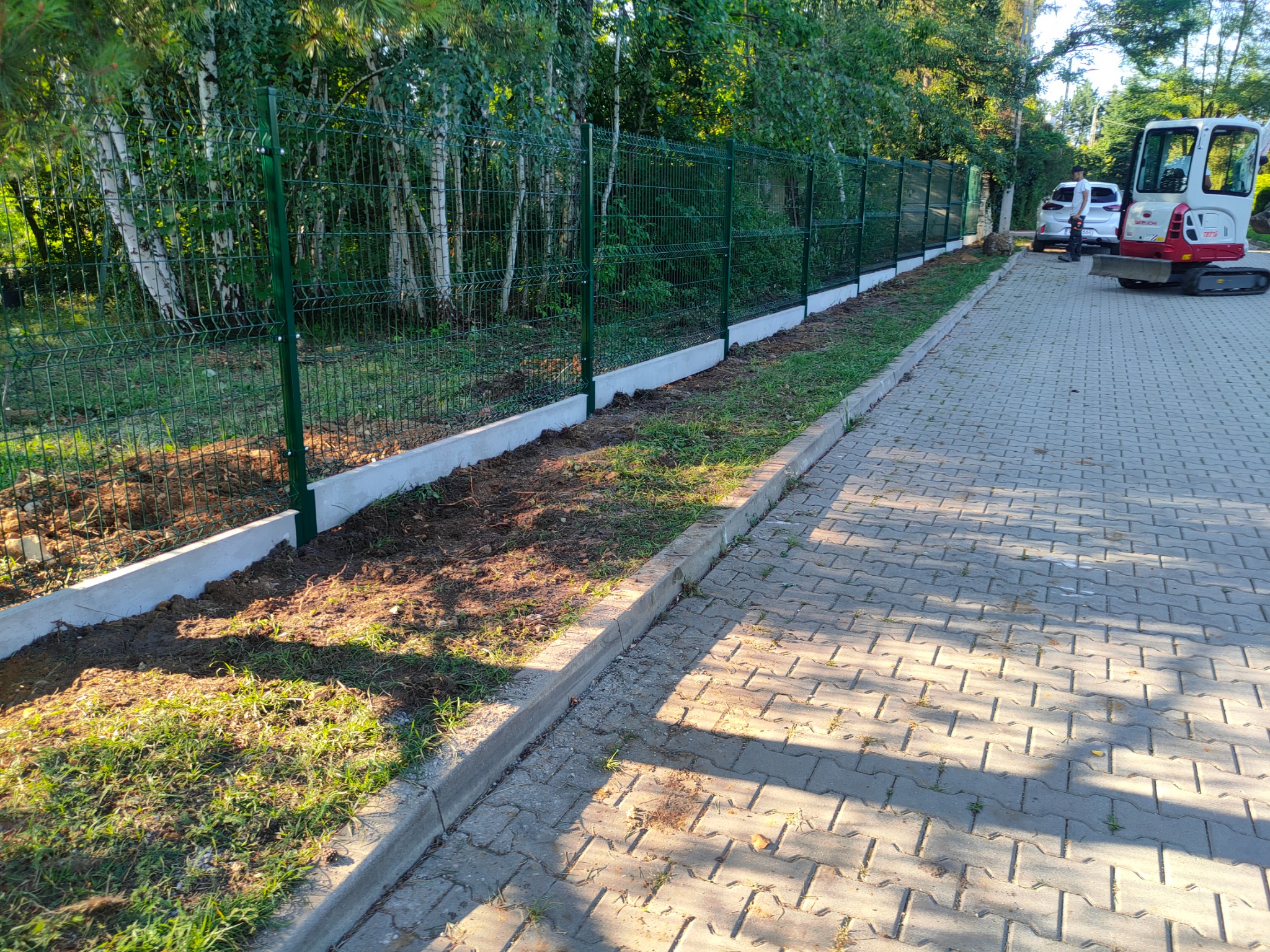 Montaż zielonego ogrodzenia panelowego na betonowej podmurówce w Porębie, widoczny mini-koparka i ułożona kostka brukowa.
